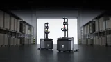 Two PUDU MP2000 autonomous pallet trucks side by side in a warehouse aisle between pallet racking, with a bright open roller door behind