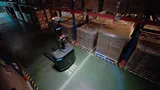 Overhead view of a PUDU MP2000 in a racked warehouse aisle beside wrapped pallets, with a 2 m aisle-width marking projected on the floor